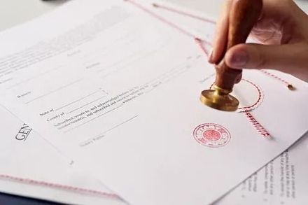 Una mano estampa un sello rojo en documentos oficiales con un hilo rojo, lo que sugiere certificación o aprobación.