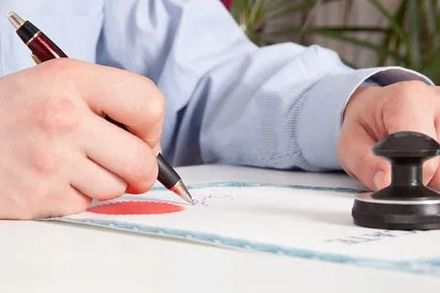 Persona firmando documento con bolígrafo, junto a un sello en papel blanco.