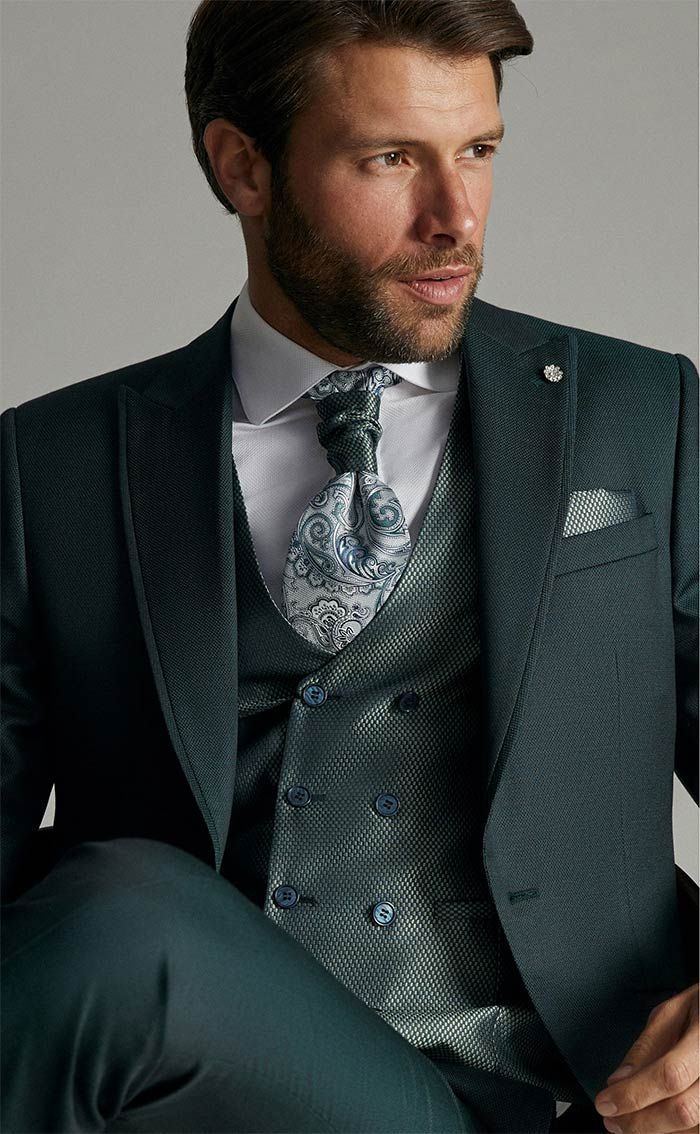 Home amb un vestit de color verd fosc, una armilla estampada i una corbata, assegut davant d’un fons neutre.