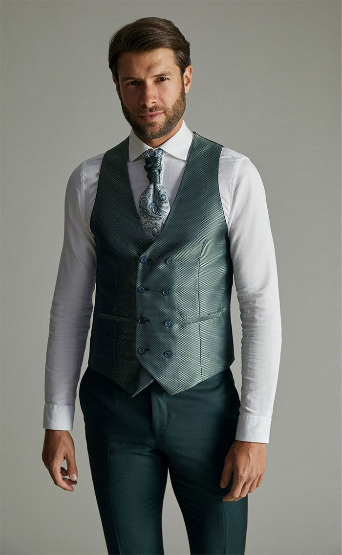 Home amb camisa blanca, armilla grisa estampada i corbata, dret davant d’un fons gris llis.