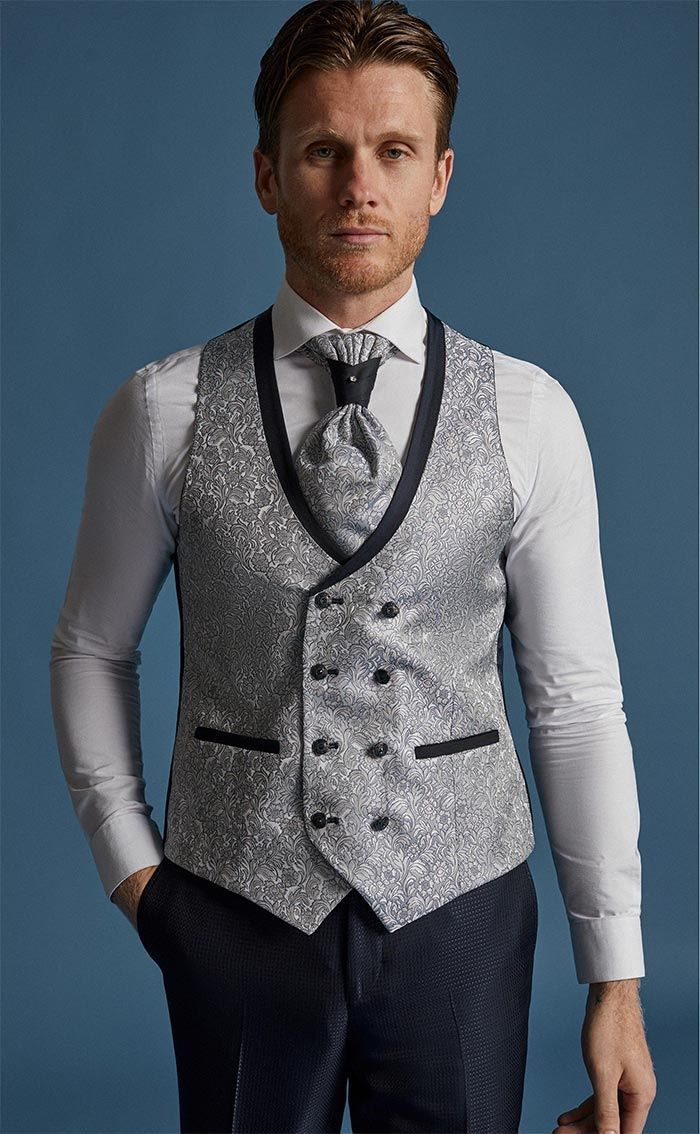 Un home amb una armilla grisa estampada sobre una camisa blanca i corbata, dret davant d’un fons blau.