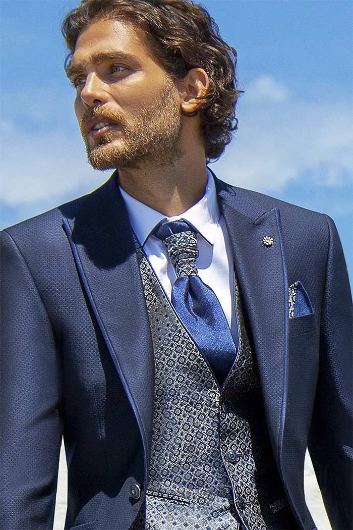 Home amb vestit blau marí i armilla estampada, mirant cap a l’esquerra a l’aire lliure, amb el cel blau de fons