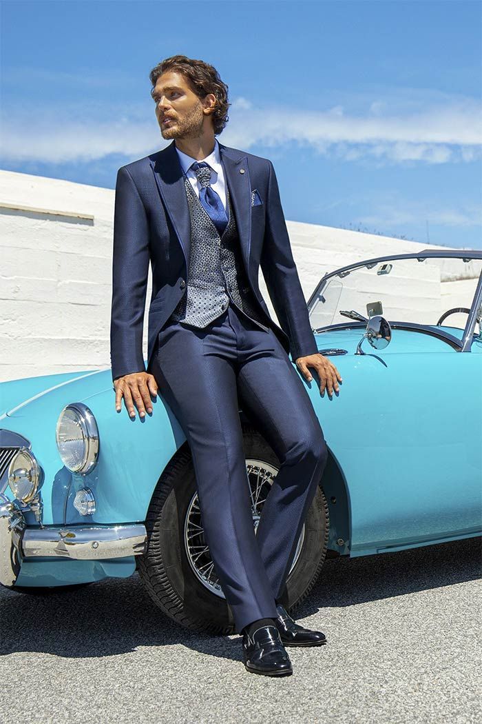 Home amb vestit blau marí recolzat en un cotxe d’època blau clar sota un cel assolellat