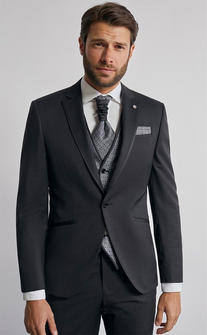 Home amb un vestit fosc, corbata estampada i mocador de butxaca, dret davant d’un fons gris clar.
