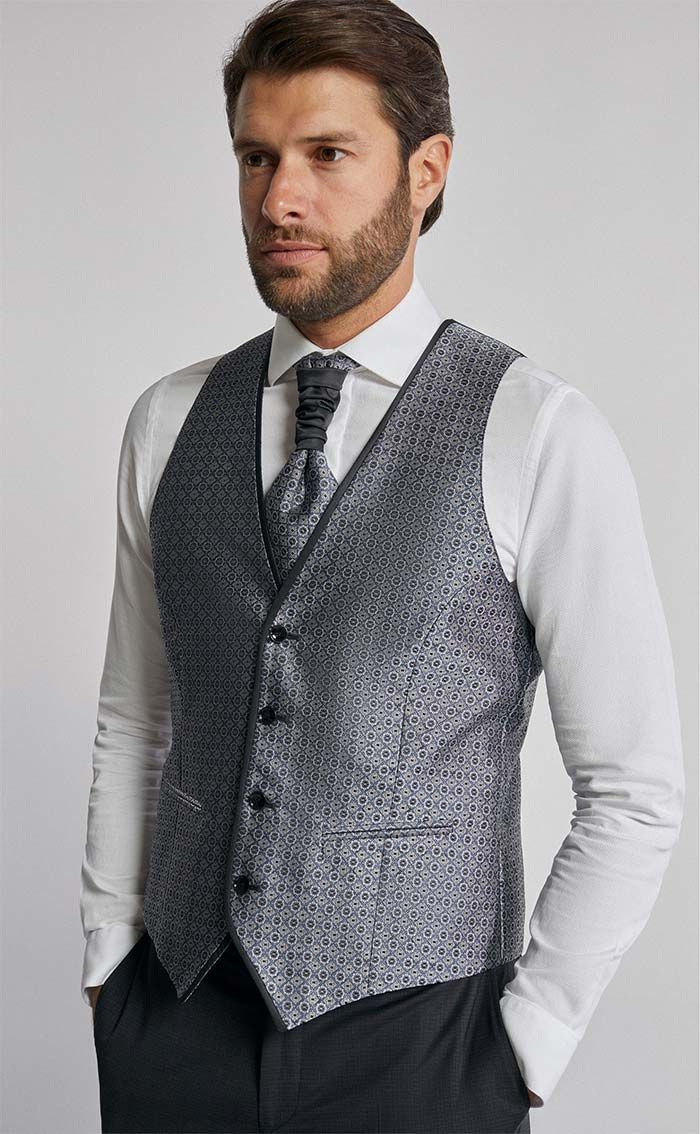 Home amb armilla grisa estampada, corbata i camisa blanca, dret amb les mans a les butxaques davant d’un fons gris