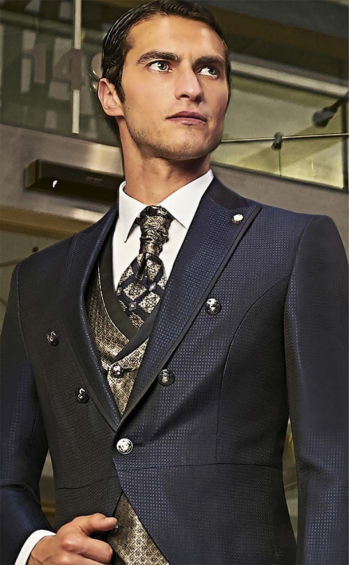 Home amb un vestit fosc de textura marcada i una corbata estampada, dret a l’interior i mirant cap a un costat.