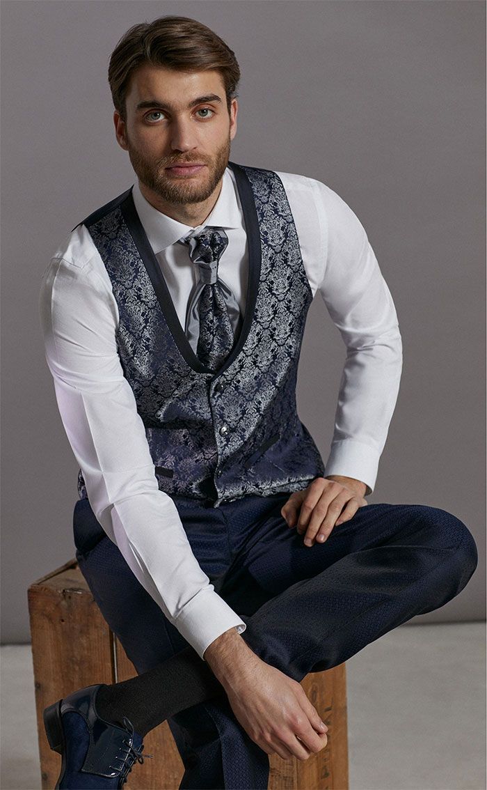 Home assegut en una caixa de fusta, amb una camisa blanca, una armilla estampada i una corbata, davant d’un fons d’estudi gris