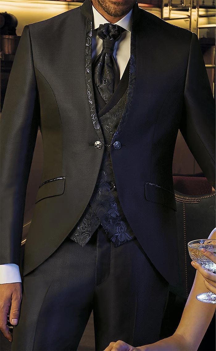 Persona amb vestit fosc, armilla i corbata estampada, dempeus al costat d’una taula amb cristalleria.
