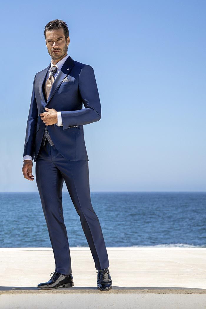 Home amb vestit blau marí posant en una terrassa davant del mar, amb l’oceà i el cel blau al darrere