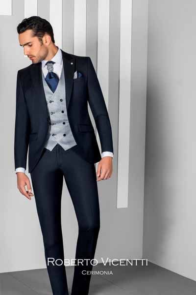 Home amb vestit blau marí, armilla grisa i corbata blava, dret davant d’un fons gris ratllat