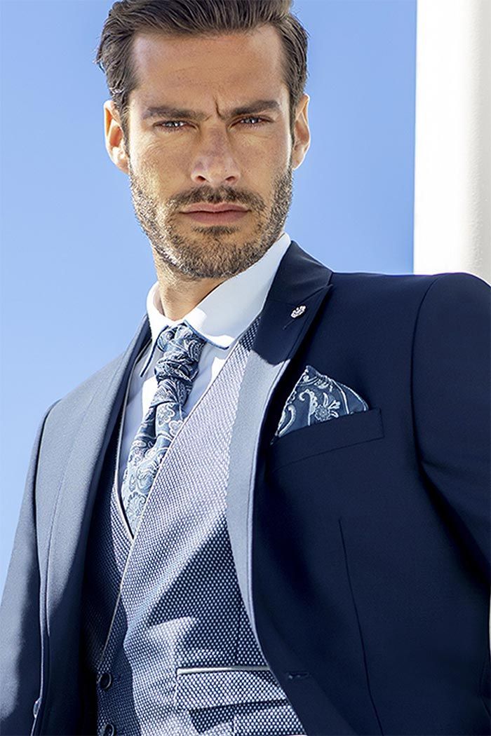 Home amb vestit blau marí i armilla estampada posant davant d’un fons blau i blanc