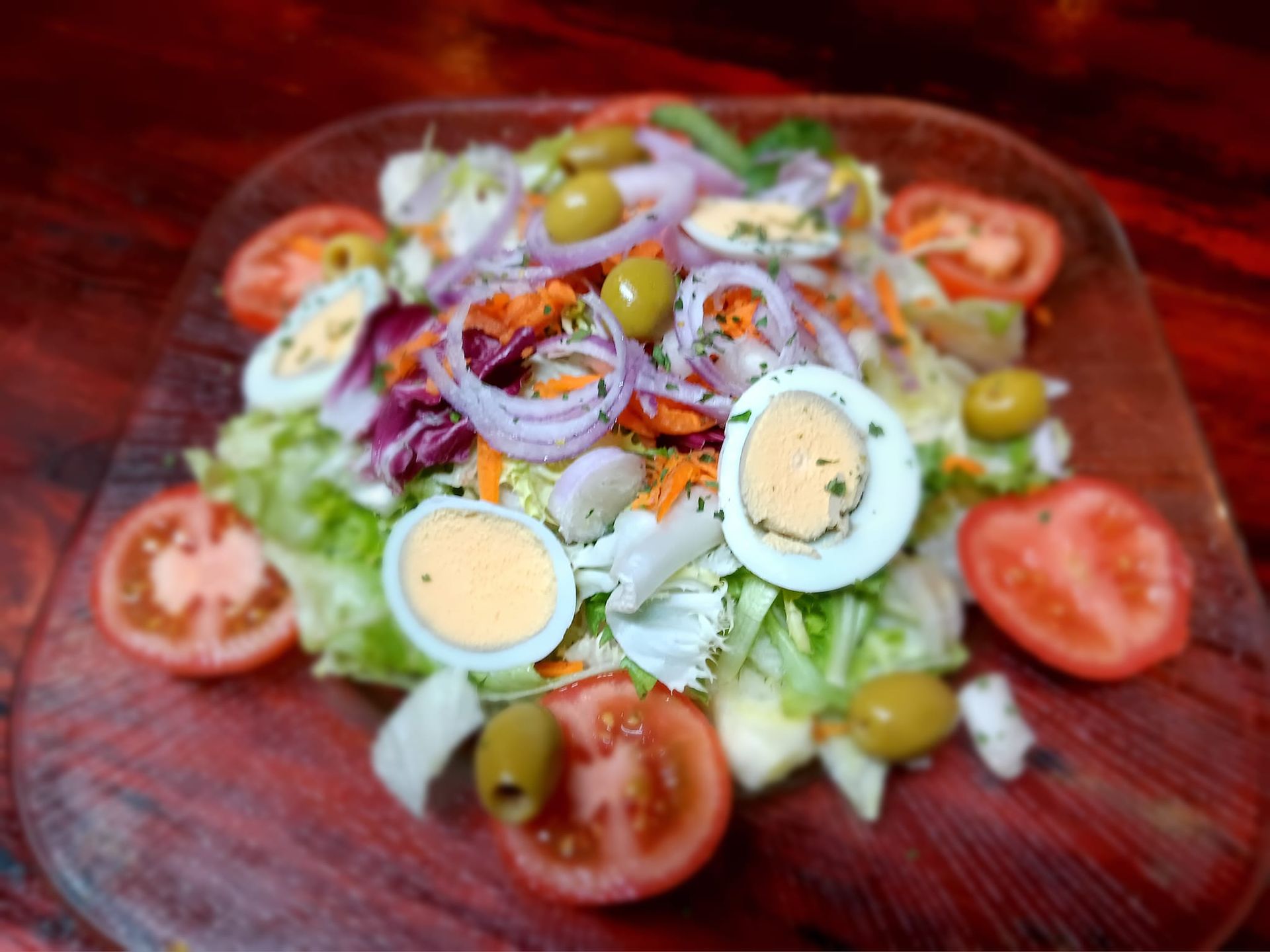 Una ensalada con tomates, huevos, aceitunas y cebollas en un plato de vidrio sobre una mesa de madera.