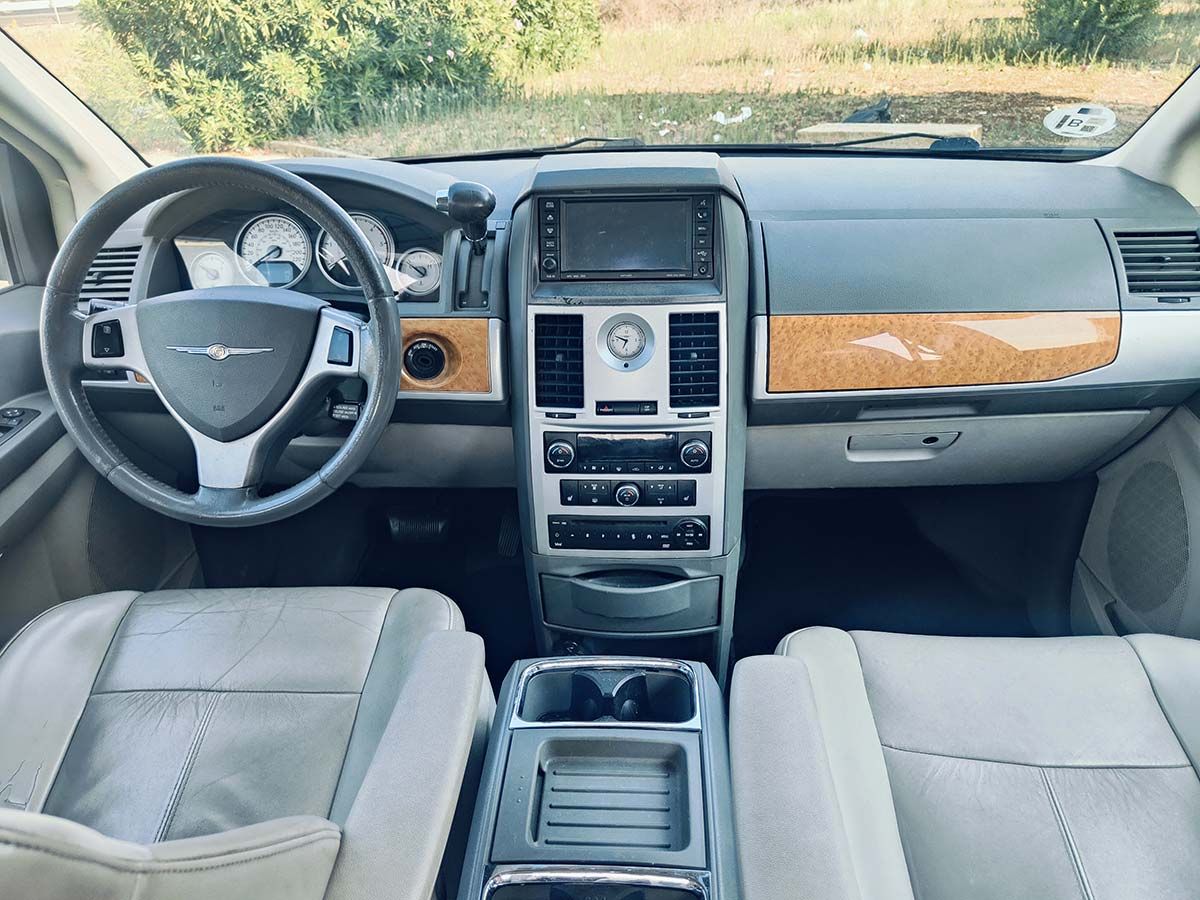 Vista interior de una minivan Chrysler gris. Tablero con molduras de madera, consola central y asientos de cuero claro.