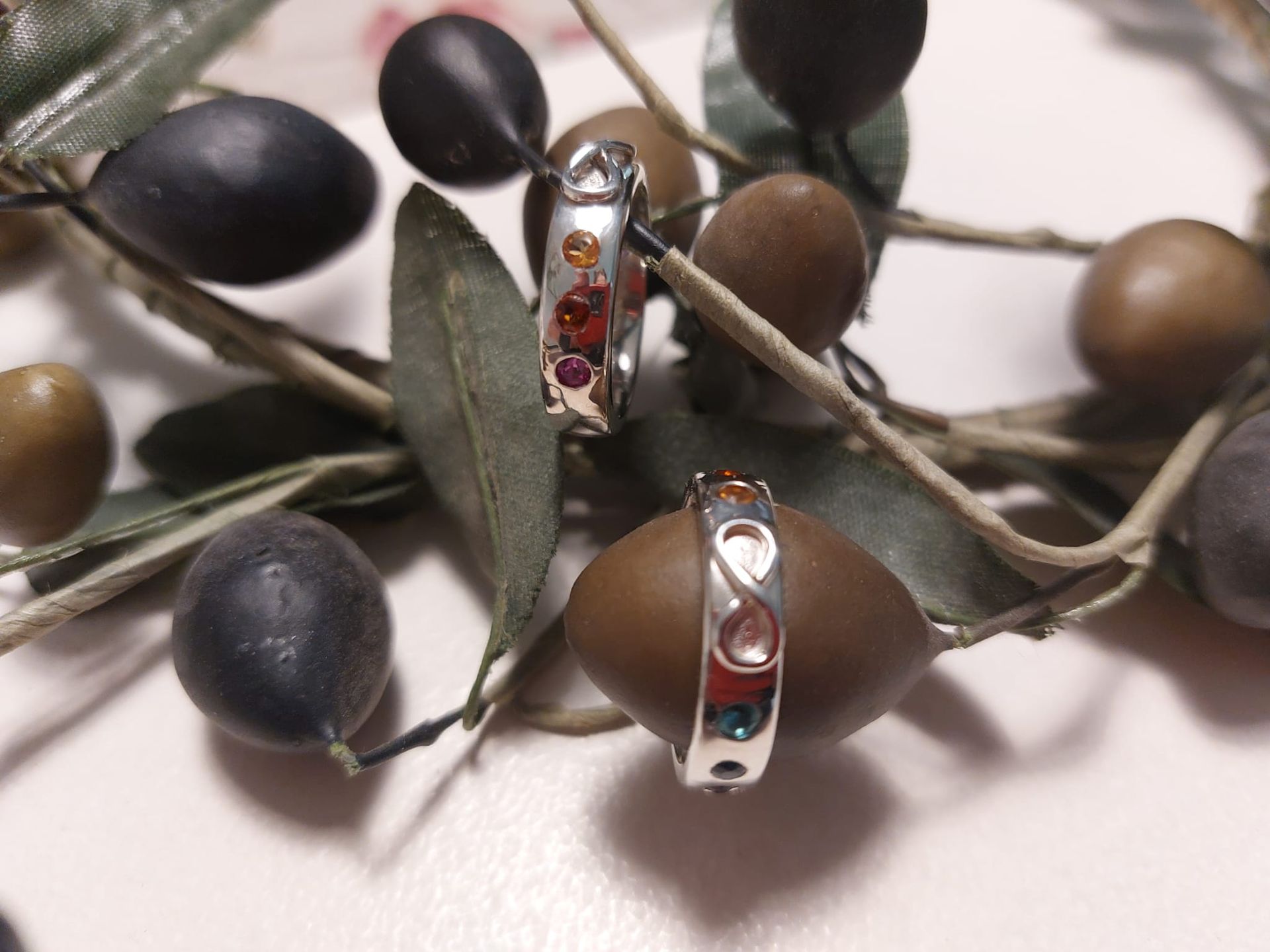 Zwei silberne Ringe mit bunten Edelsteinen, präsentiert auf einem Olivenzweig mit Oliven.