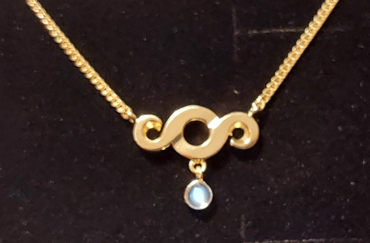 Goldene Halskette mit einem stilisierten „S“-Anhänger und einem kleinen blauen Edelstein.