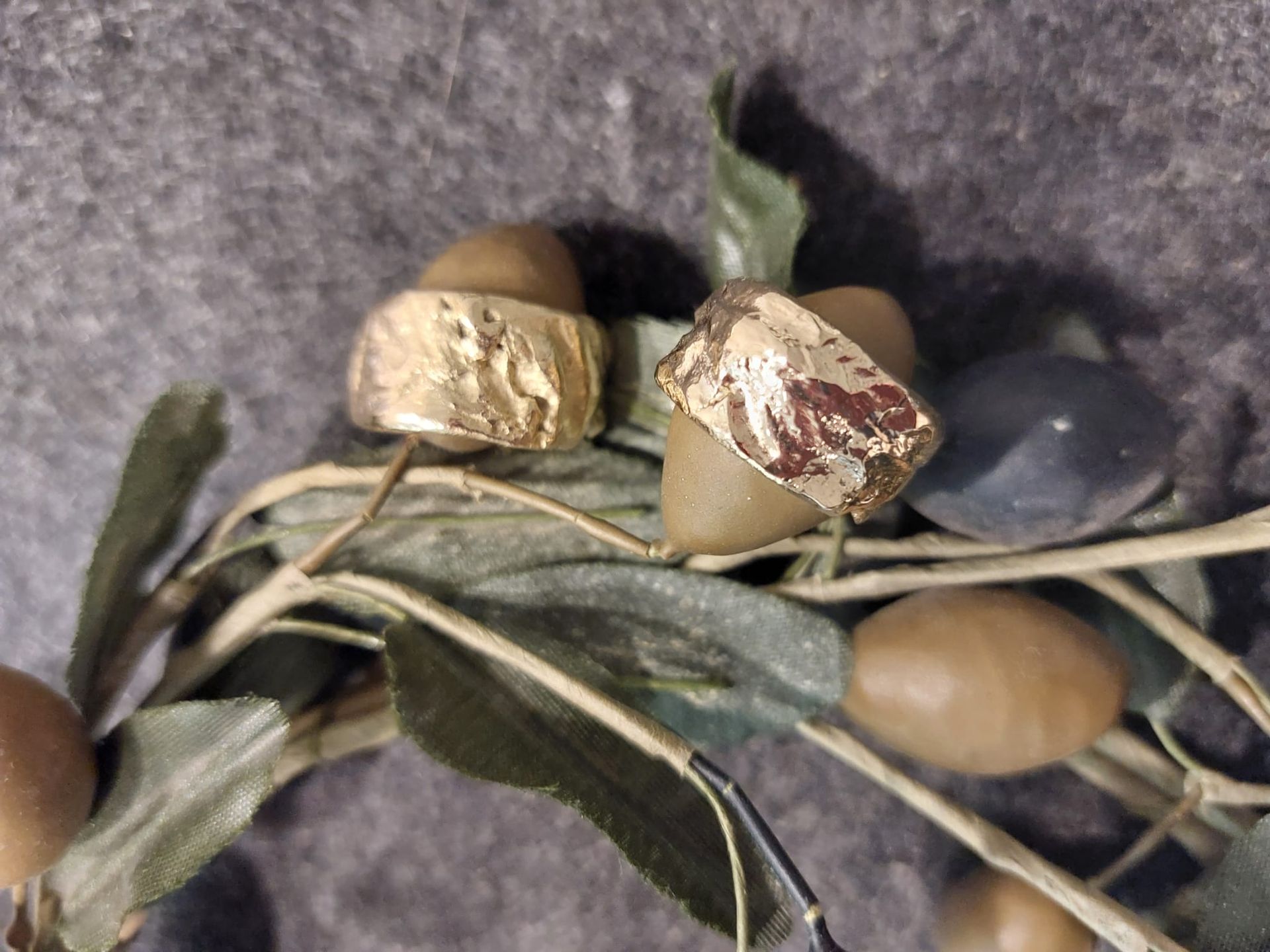 Zwei Ringe mit Goldstruktur ruhen auf einem künstlichen Olivenzweig mit grünen Blättern und olivfarbenen Früchten.