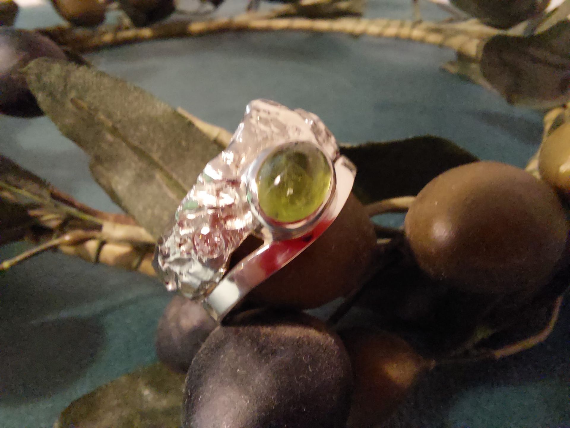 Silberring mit einem grünen Edelstein, der auf dunklen Oliven und Blättern ruht.