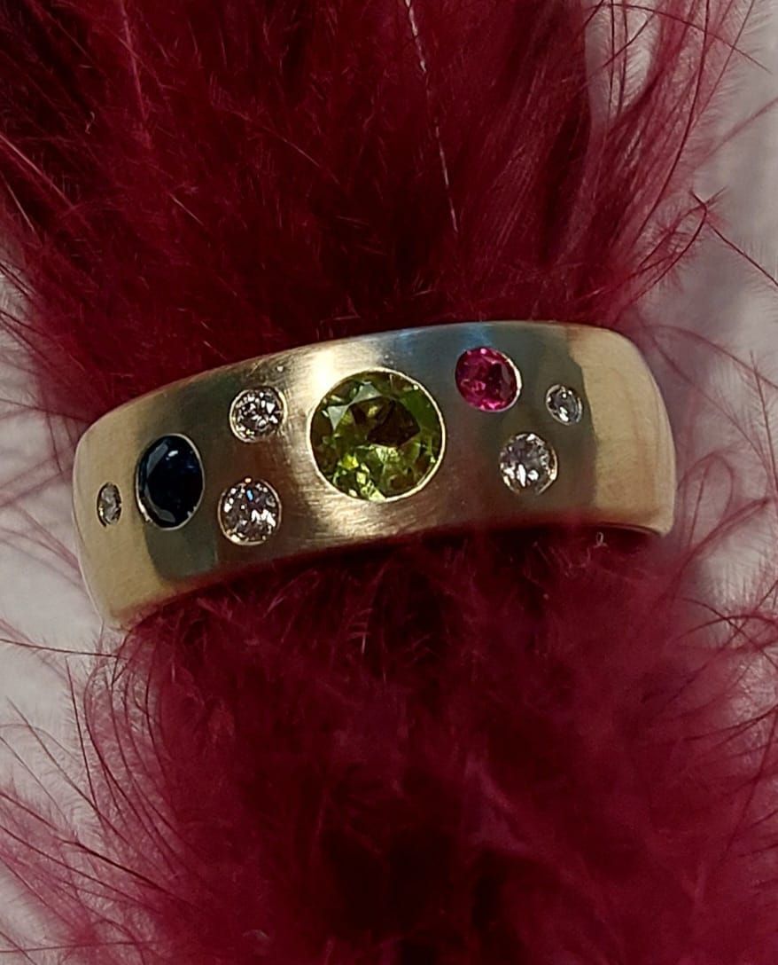 Goldring mit eingelegten Edelsteinen (blau, rosa, grün, klar), ruhend auf roten Federn.
