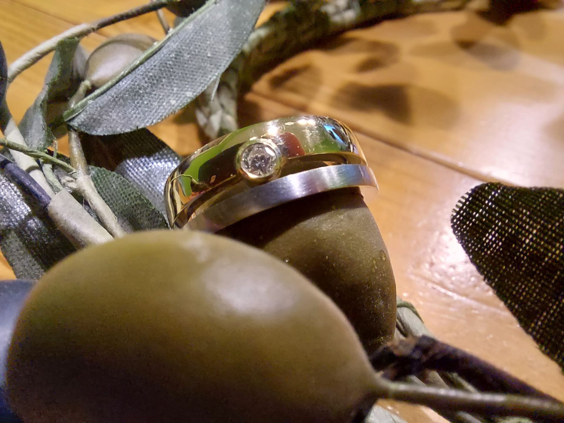 Zwei Ringe, einer davon aus Gold mit einem Diamanten, sind auf einem Silberband gestapelt und ruhen auf grünen Oliven mit Blättern auf einer Holzoberfläche.