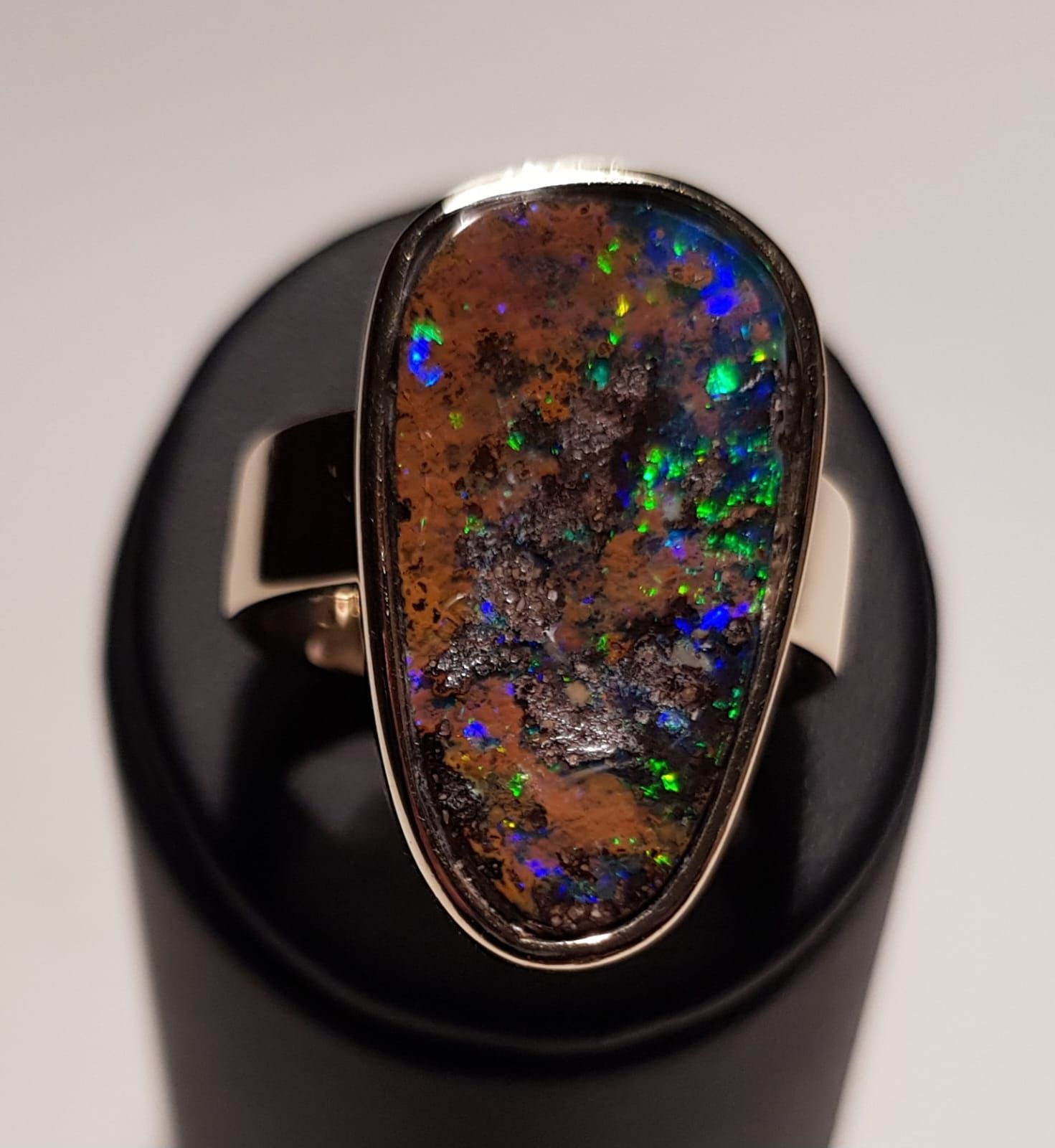Ring mit einem braun-schwarzen Boulderopal, der ein grünes, blaues und gelbes Farbspiel zeigt, gefasst in ein Goldband.