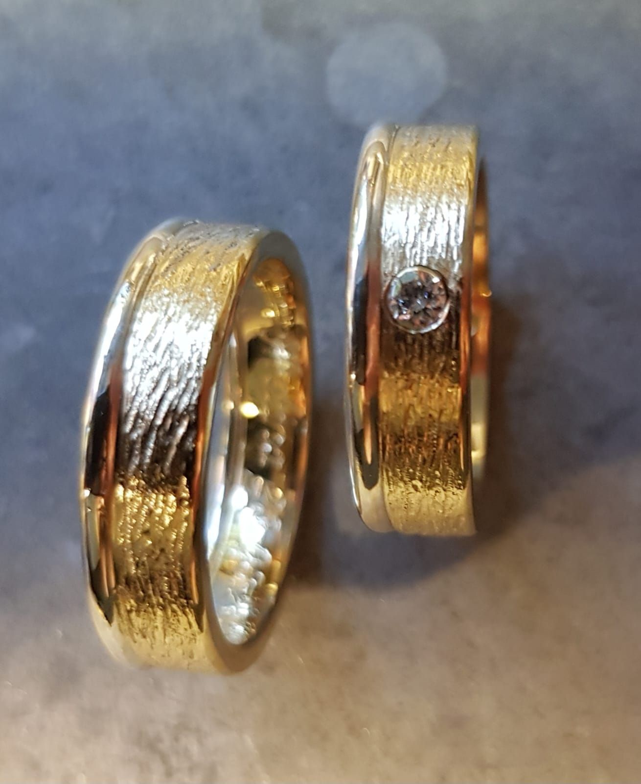 Zwei goldene Eheringe mit strukturierten Außenbändern. Ein Ring ist mit einem einzelnen runden Diamanten besetzt.