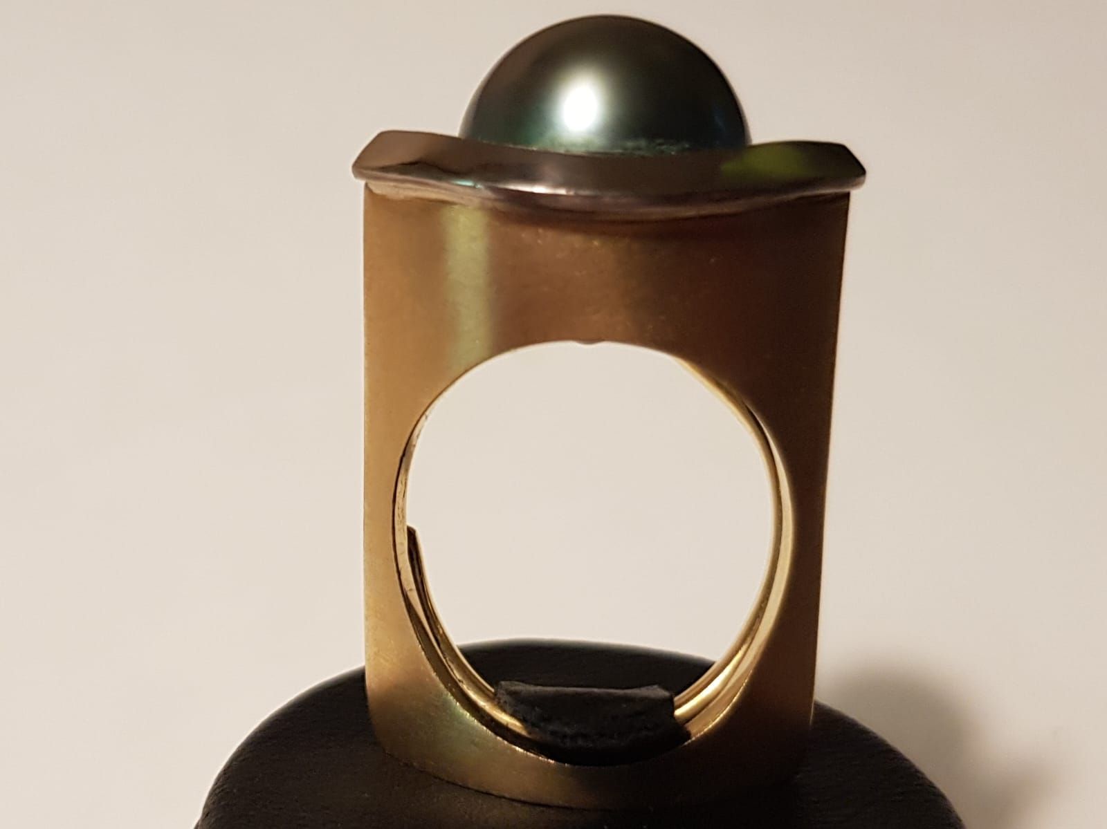 Goldfarbener Ring mit einer oben eingefassten dunklen Perle, in der Mitte befindet sich eine große kreisrunde Öffnung.