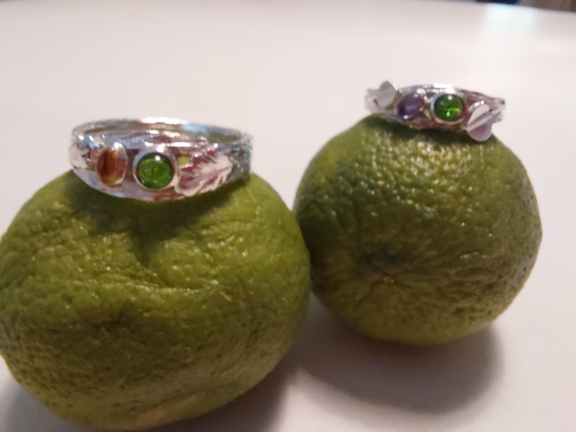 Zwei Edelsteinringe liegen auf grünen Zitrusfrüchten vor einer weißen Oberfläche.