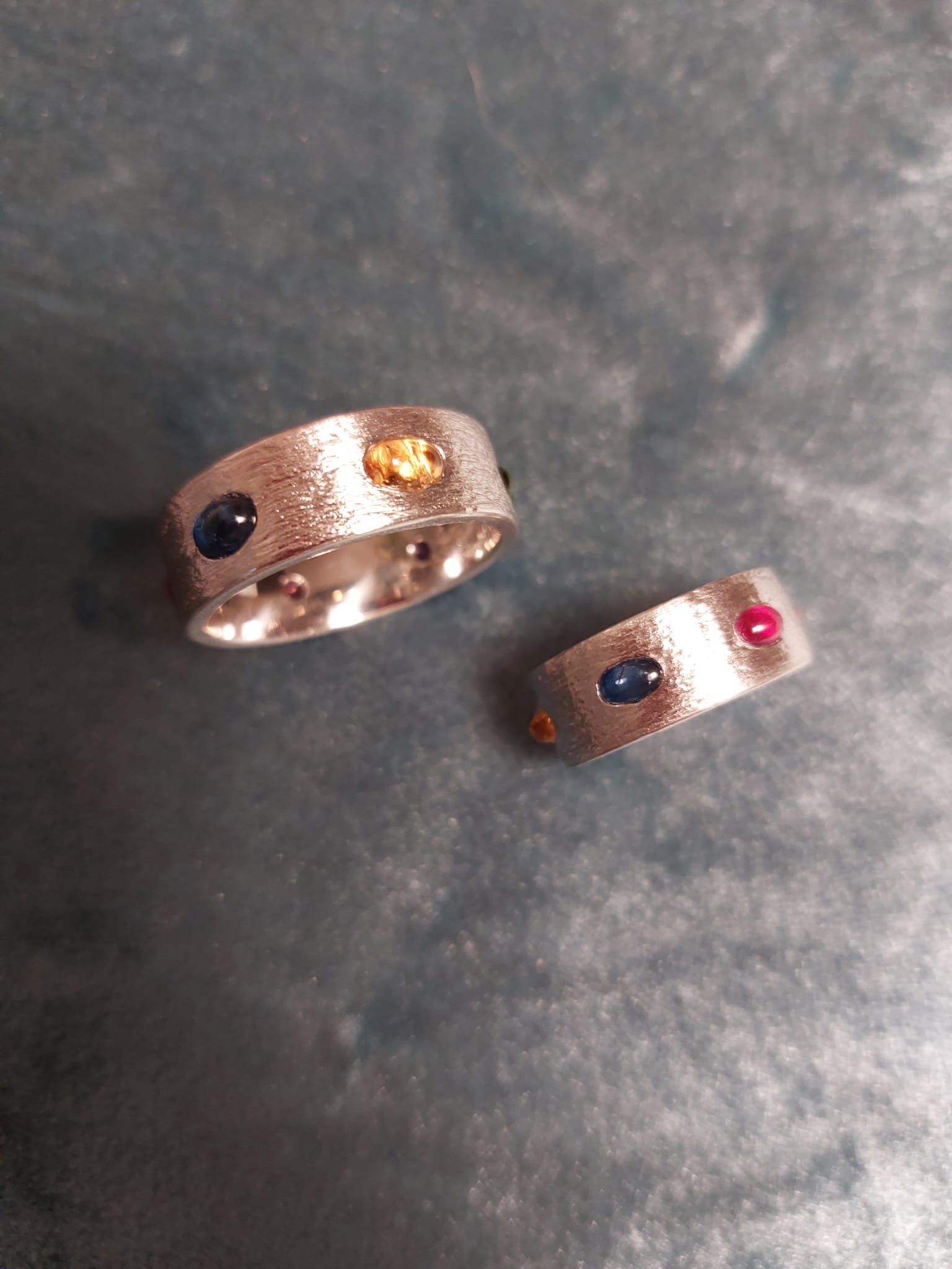 Zwei Silberringe mit eingelegten Edelsteinen auf einer strukturierten Oberfläche; ein Ring hat blaue und gelbe Steine, der andere hat blaue und rote Steine.