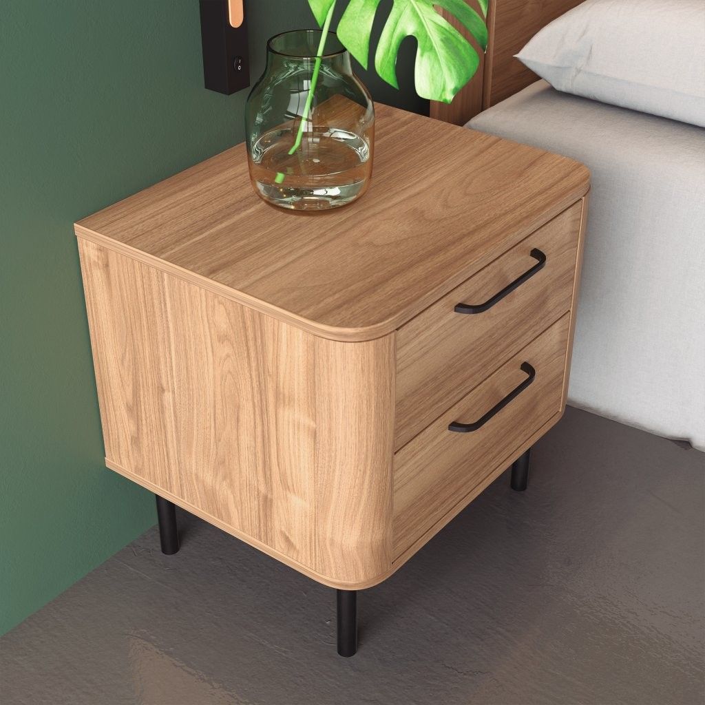 Mesita de noche de madera con dos cajones y un jarrón, junto a una cama con una pared verde.