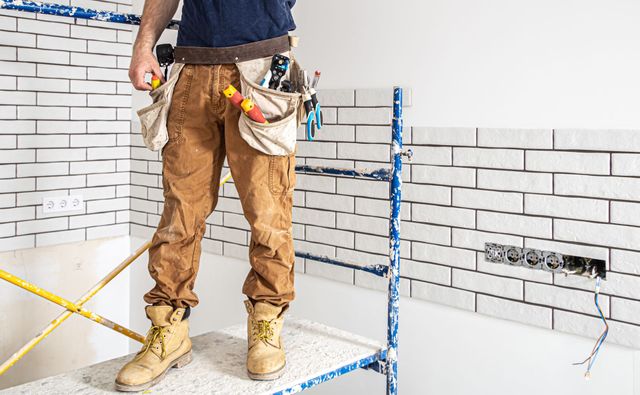 Obrero de la construcción de pie sobre una pared de azulejos, sosteniendo herramientas junto a una habitación parcialmente alicatada con andamios.