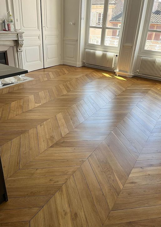 Parquet à chevrons en bois baigné de lumière dans une pièce lumineuse aux grandes fenêtres et aux portes blanches à panneaux