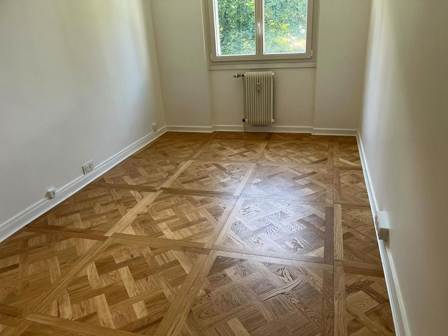 Pièce vide avec parquet en chevrons, murs blancs et fenêtre avec un radiateur en dessous.