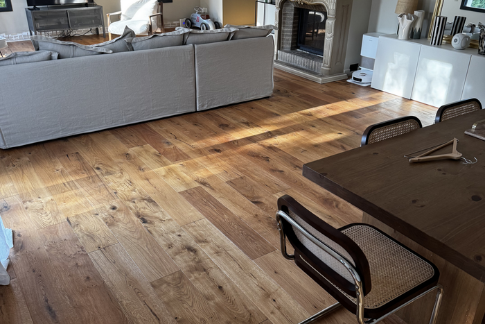 Salon lumineux avec parquet, canapé d’angle gris et table à manger en bois avec des chaises.