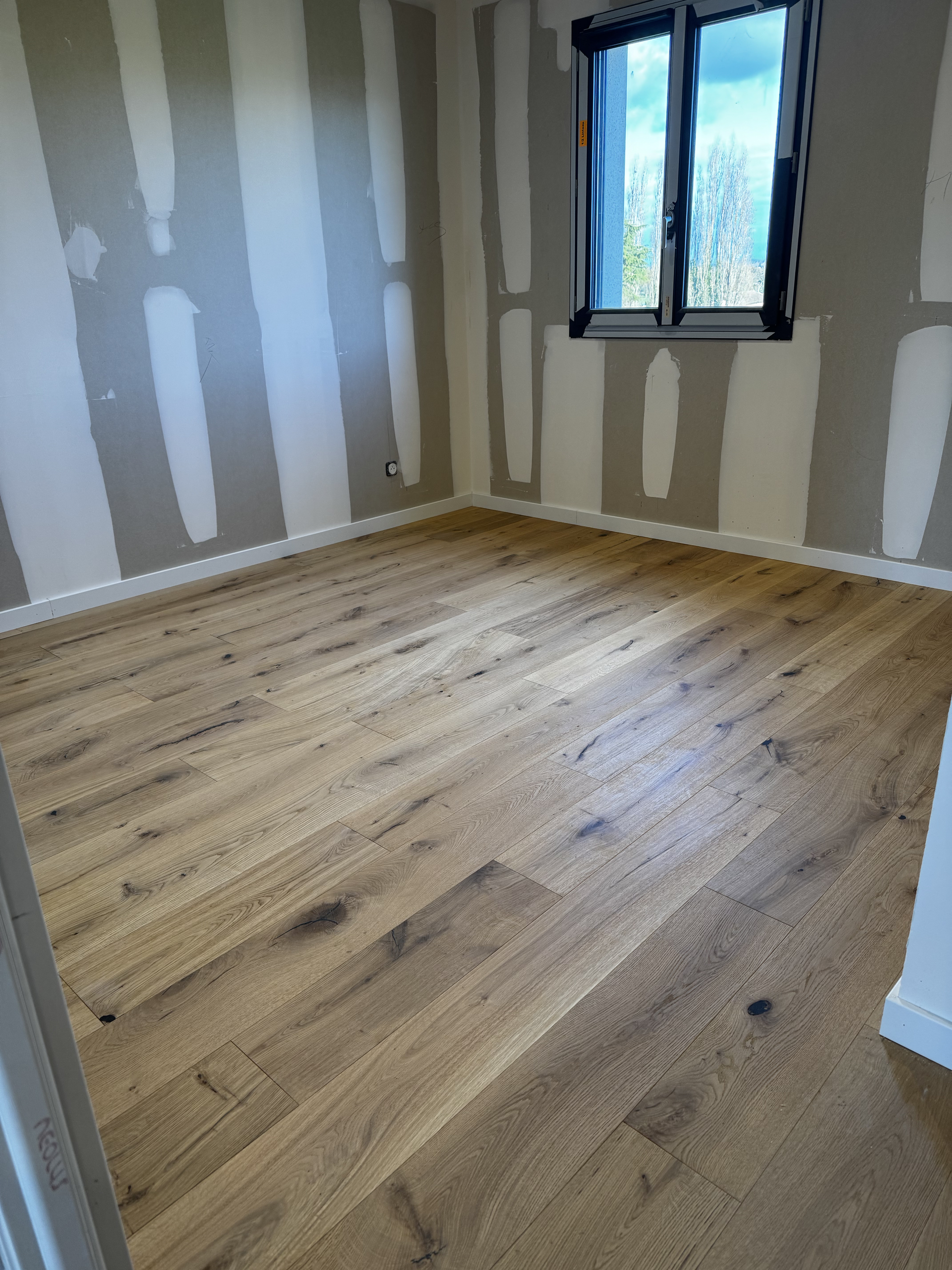 Pièce vide avec des plaques de plâtre non finies, un parquet clair et une fenêtre laissant entrer la lumière du jour.
