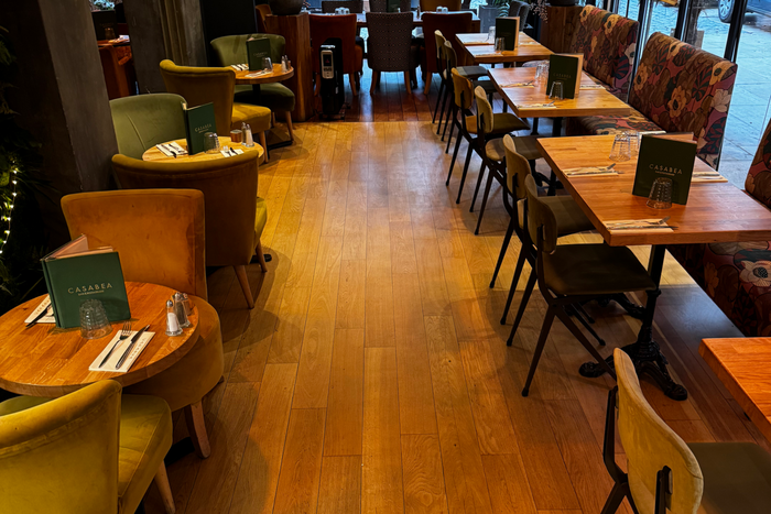 Salon vide avec un parquet clair, un canapé gris et deux petites tables.