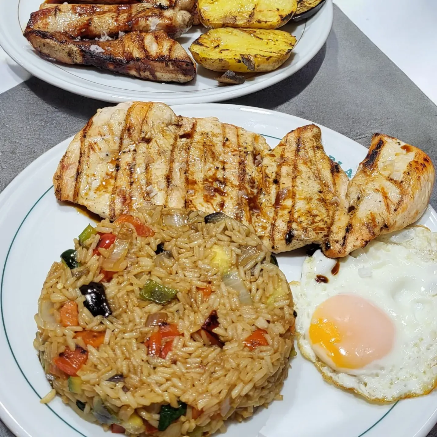 Dos platos de pollo a la parrilla, uno con arroz con verduras y un huevo frito, y el otro con patatas asadas.