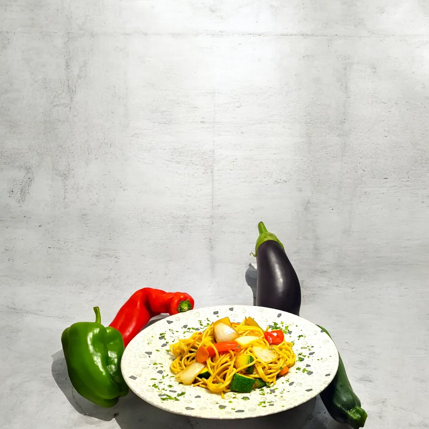 Un plato de pasta con verduras reposa sobre una superficie gris, rodeado de un pimiento verde, un pimiento rojo, una berenjena y un calabacín.