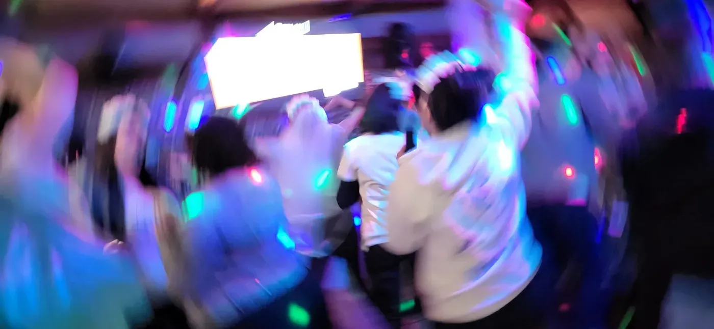 Una escena enérgica y con efecto de desenfoque de movimiento de personas bailando en una fiesta con luces de colores y una pantalla blanca brillante.