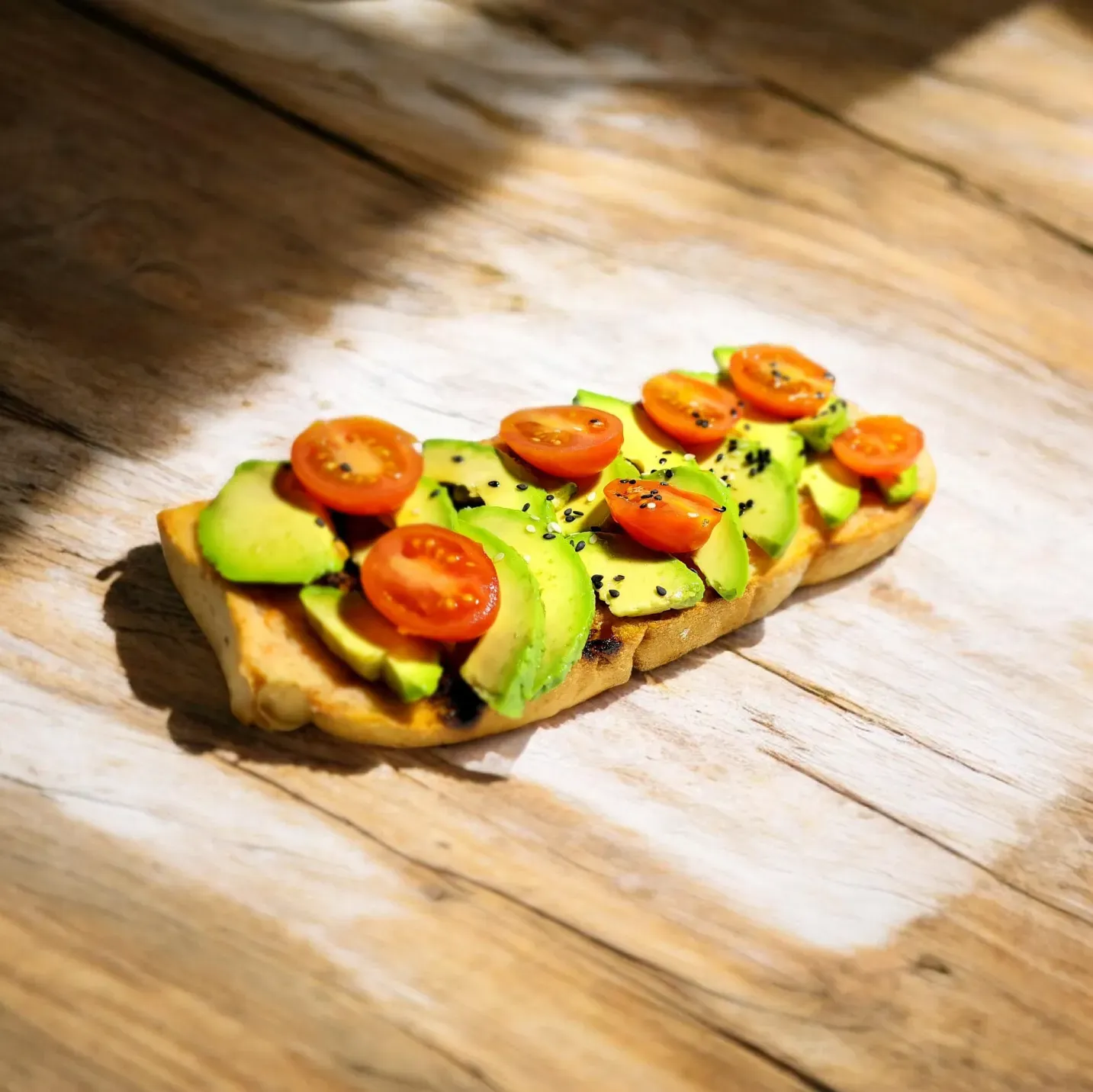 Tostada de aguacate con tomates cherry y semillas de sésamo negro sobre una mesa rústica de madera, bajo la luz del sol.