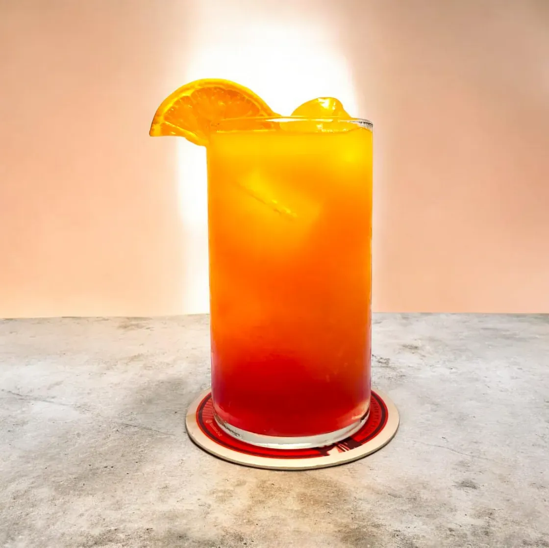 Un cóctel Tequila Sunrise de color naranja brillante con el fondo en degradado rojo y una rodaja de naranja como guarnición, servido sobre un posavasos redondo.