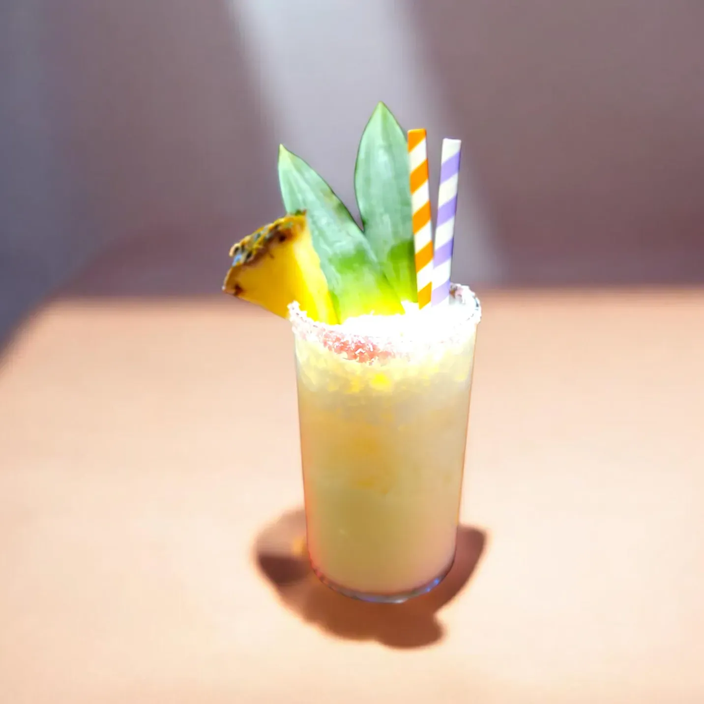 Un cóctel cremoso de color amarillo servido en un vaso con una rodaja de piña, hojas y dos pajitas a rayas, sobre un fondo color melocotón.