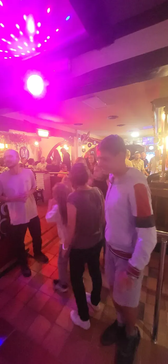 Dos personas cantan karaoke en un escenario dentro de un bar con poca luz, iluminado por coloridas