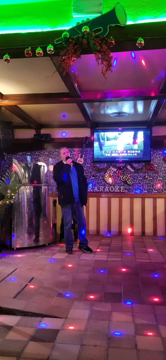Dos personas están sentadas en una mesita en un pub con paredes rojas, bebiendo cerveza y vino, y sonriendo a la cámara.