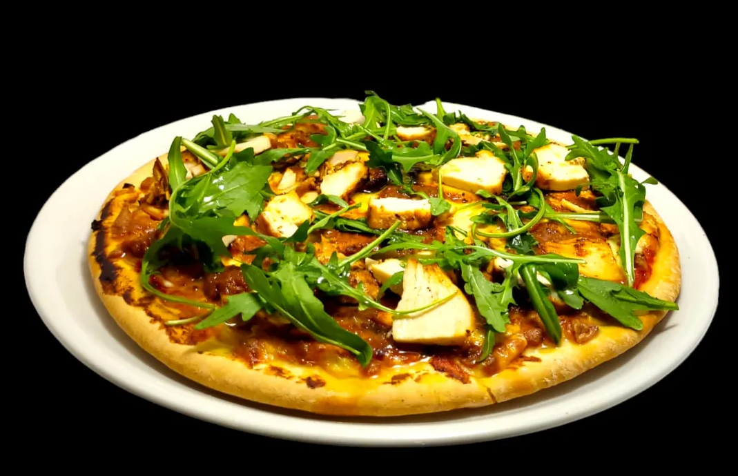 Una pizza cubierta con trozos de pollo y rúcula fresca sobre un plato blanco con fondo negro.