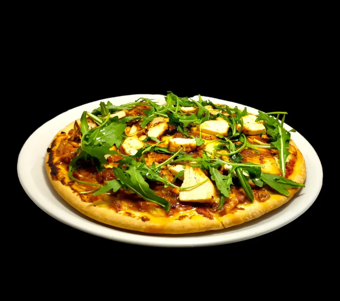Una pizza cubierta con trozos de pollo y rúcula fresca sobre un plato blanco con fondo negro.