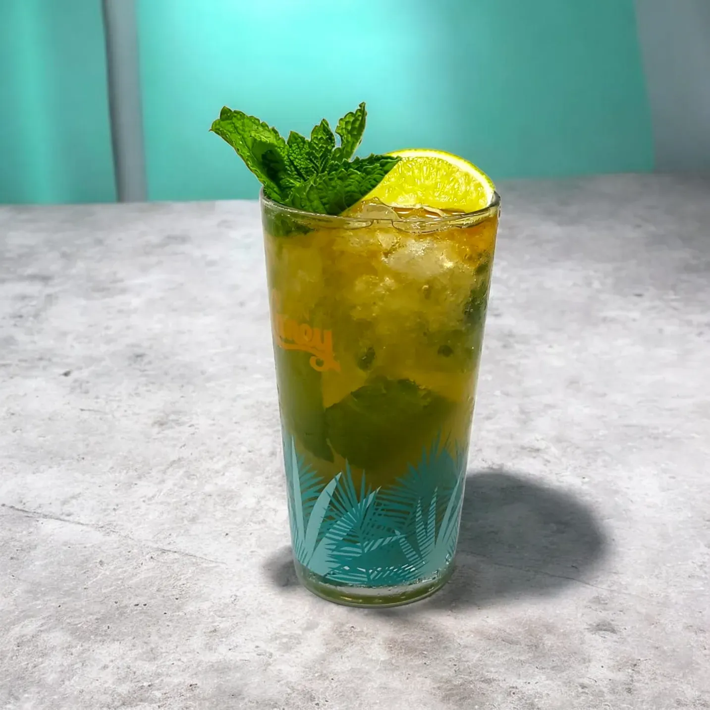 Una refrescante bebida helada servida en un vaso con estampado de palmeras, adornada con una rodaja de lima y una ramita de menta sobre una superficie gris.