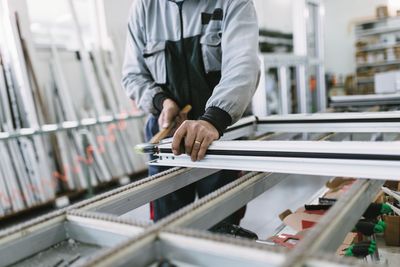 Un ouvrier mesure un cadre de fenêtre dans un atelier où des cadres et des outils sont empilés à proximité.