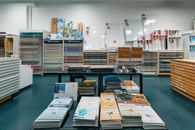 Hyvin järjestetty taidetarvikeliike, jossa on riveittäin papereita, taidetauluja ja luetteloita hyllyillä ja esittelypöydillä.