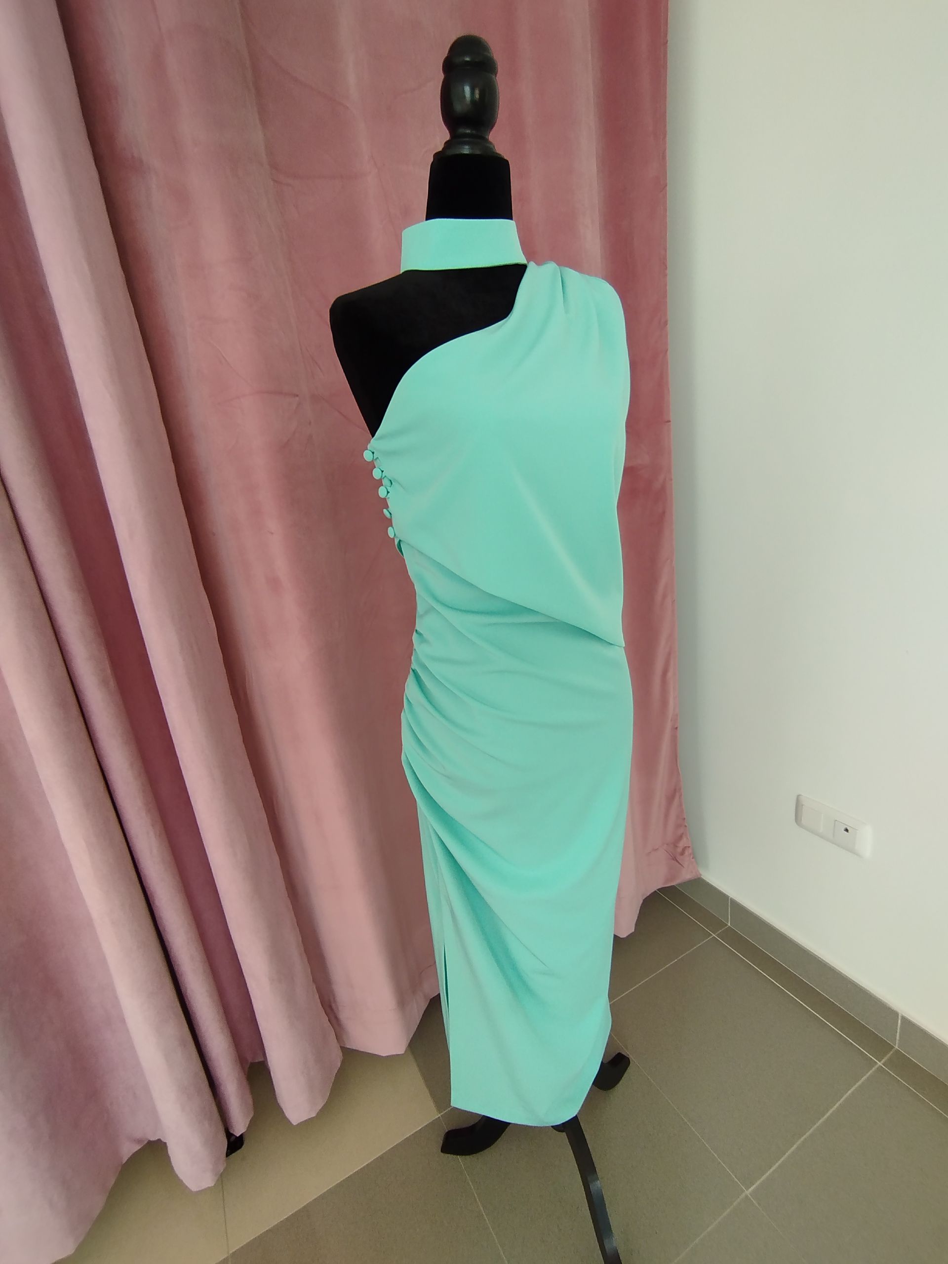 Un vestido de un solo hombro de color verde menta sobre un maniquí negro contra una cortina rosa y una pared blanca.