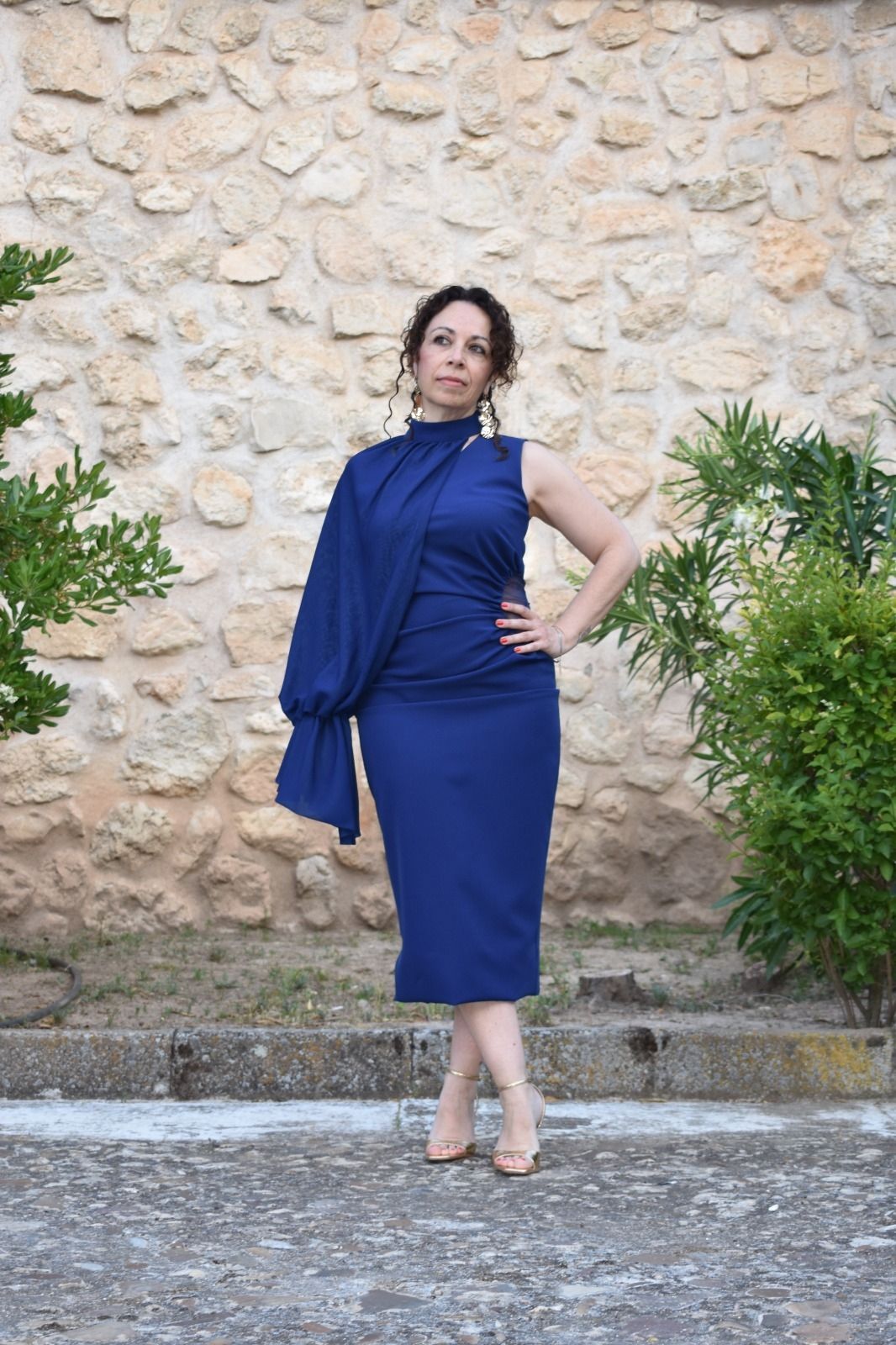 Una mujer con un vestido azul oscuro de una sola manga posa frente a un muro de piedra.